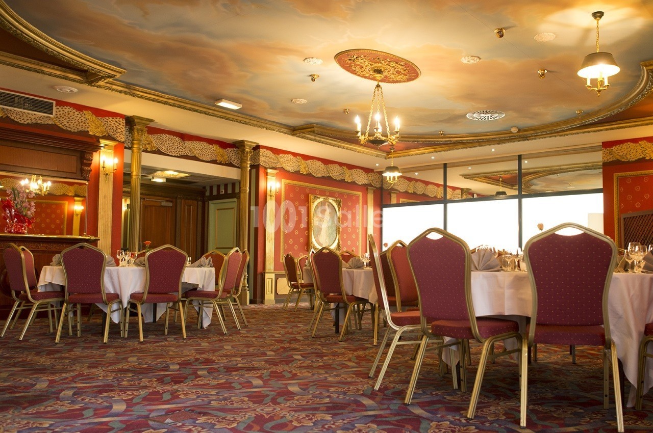 Salle de réception élégante avec tables rondes dressées, chaises rouges et plafond décoré de fresques.