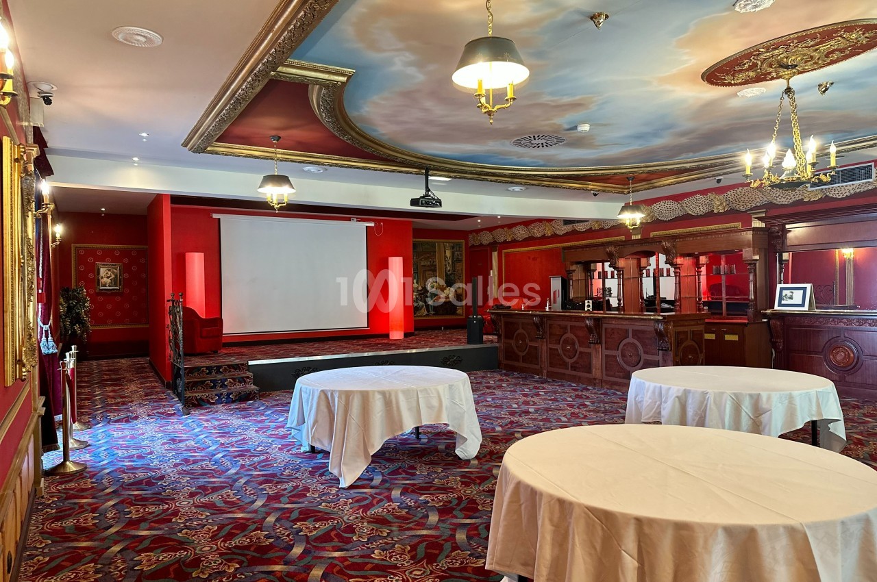 Salle de réception décorée de rouge avec tables rondes, scène équipée d'un écran et plafond peint de motifs artistiques.