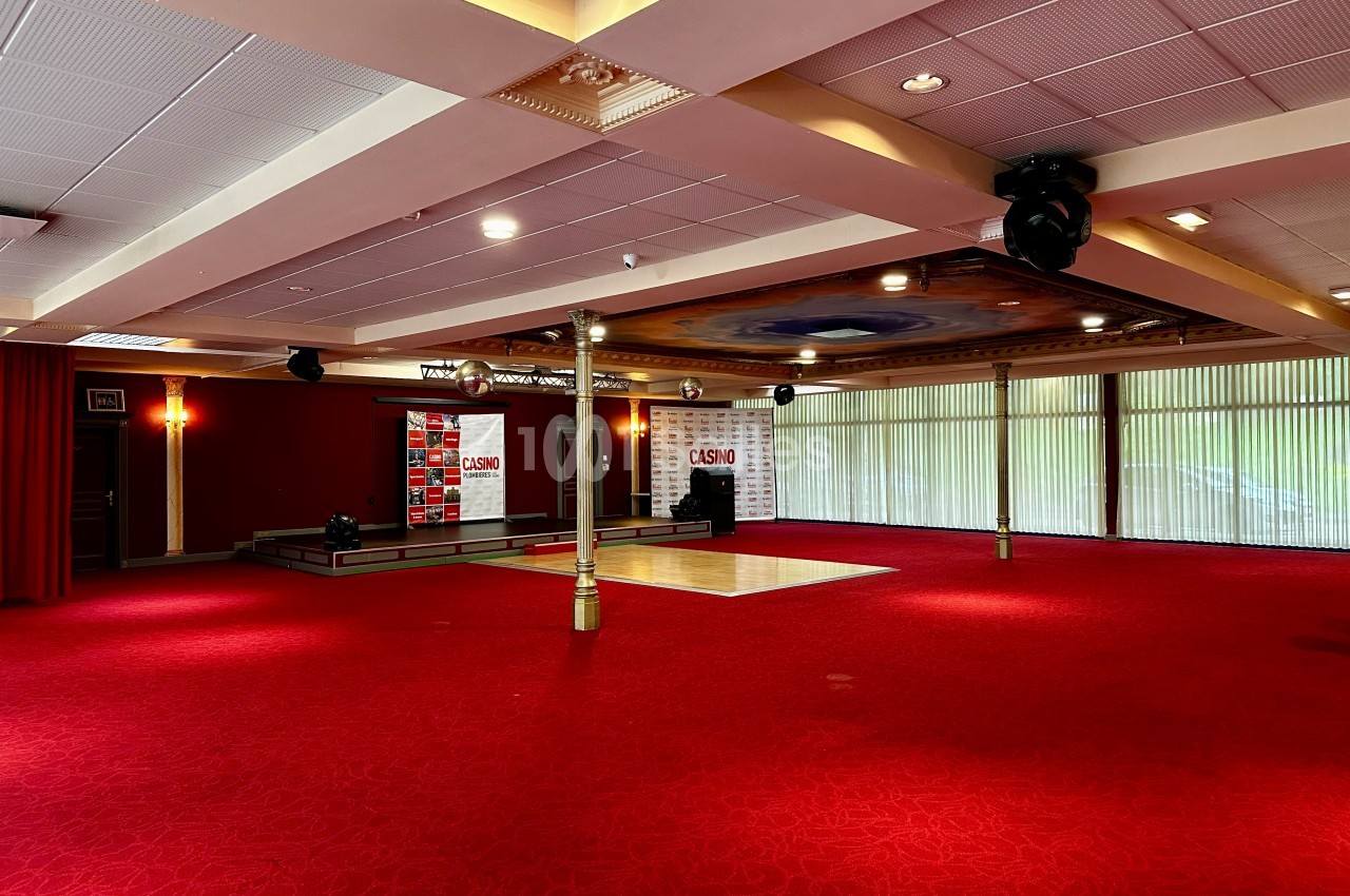 Salle spacieuse avec moquette rouge, petite scène, rideaux rouges et grandes baies vitrées laissant entrer la lumière.