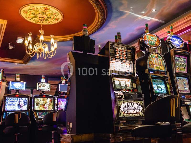 Salle de jeux avec des machines à sous alignées sous un plafond décoré et un lustre suspendu.