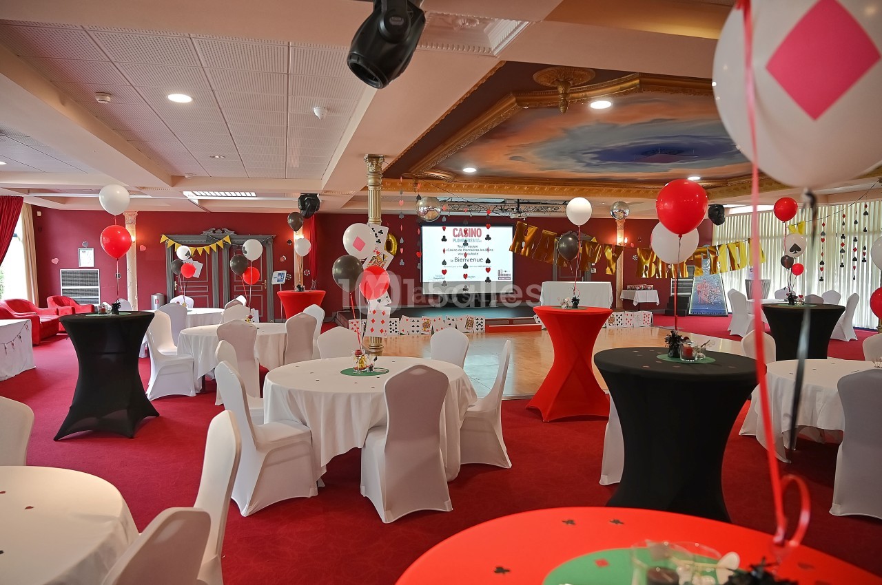 Salle décorée sur le thème du casino avec tables rondes, nappes blanches, ballons et écran de projection.