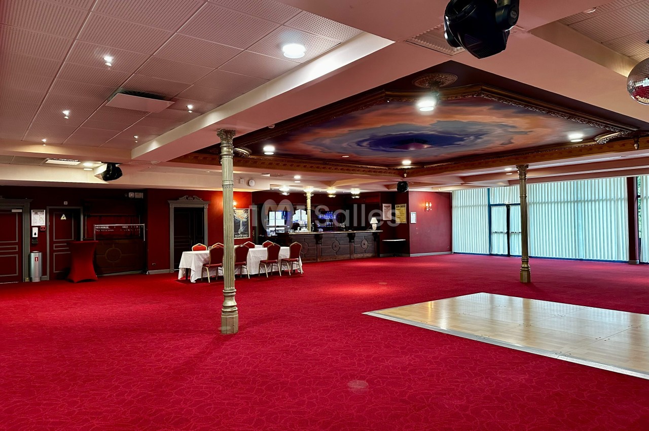 Salle spacieuse avec tapis rouge, piste de danse en bois, tables dressées et bar en arrière-plan.