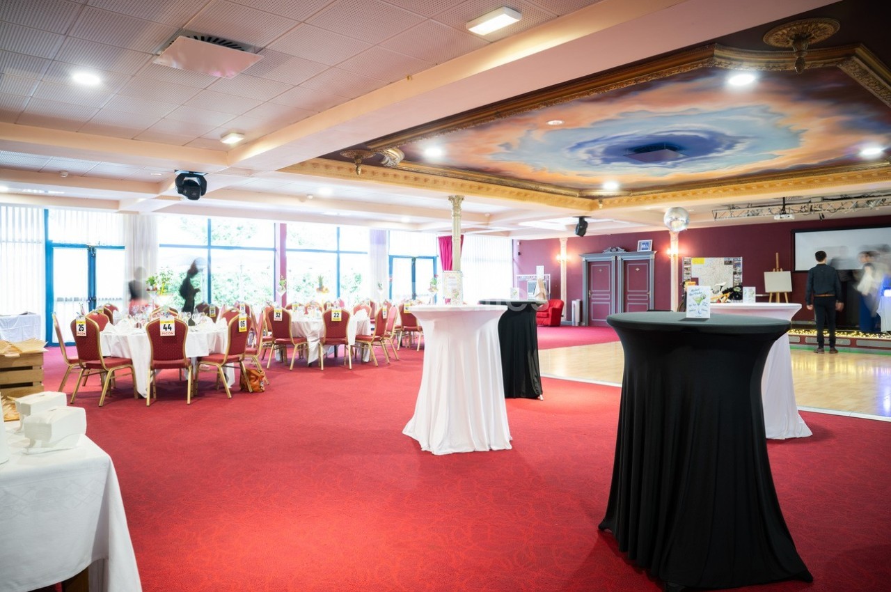 Salle de réception avec tables dressées, nappes blanches, chaises rouges et piste de danse sous un plafond décoré.