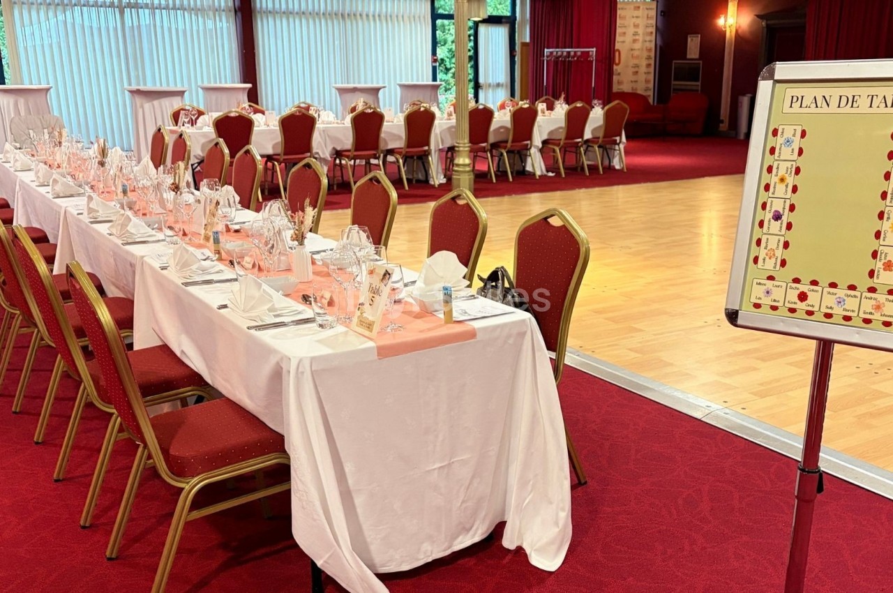Salle de réception avec tables dressées, chaises rouges et un plan de table affiché près d'une piste de danse.