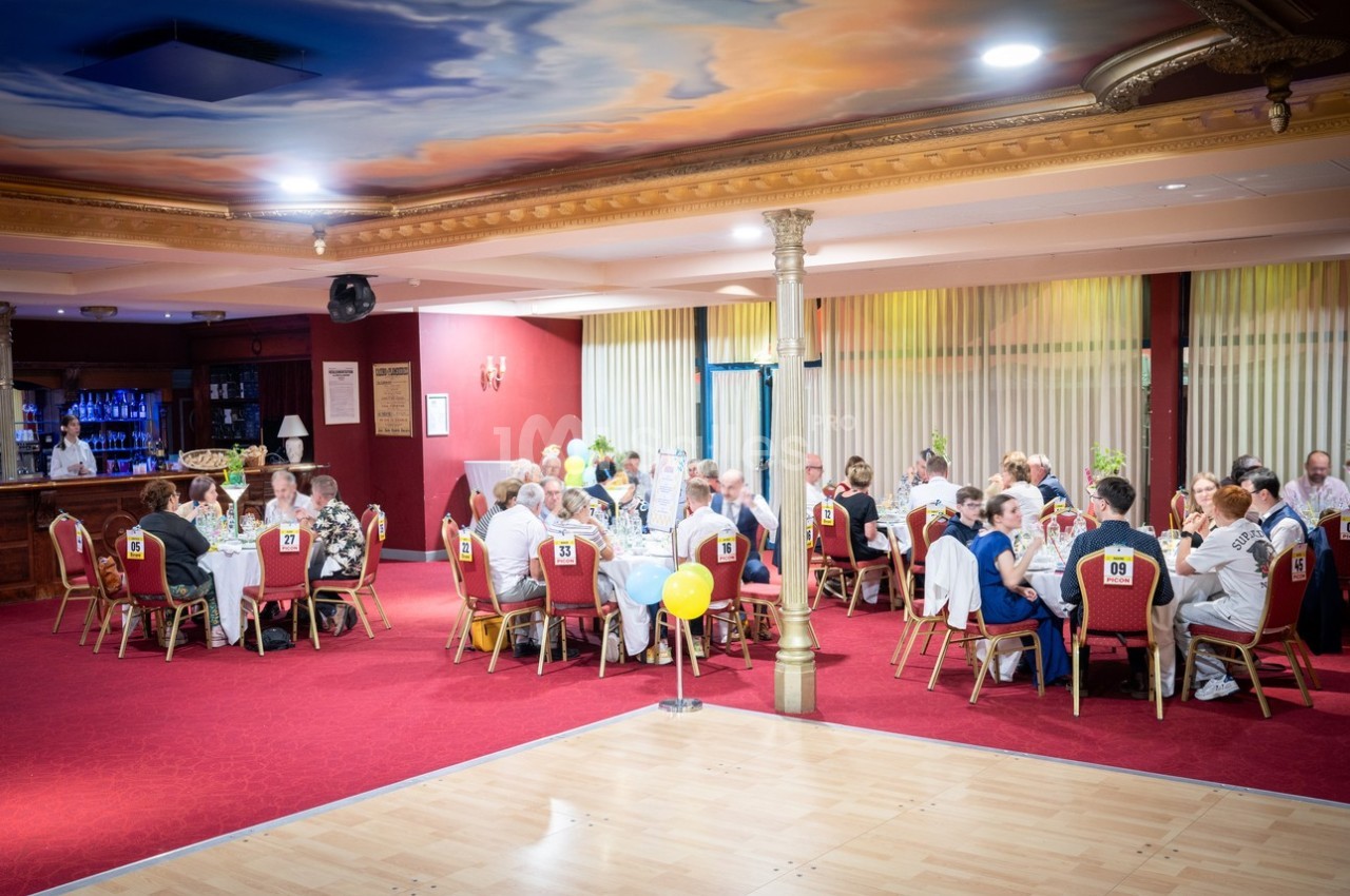 Des convives assis autour de tables rondes dans une salle de réception décorée, avec un plafond peint et un sol en parquet.