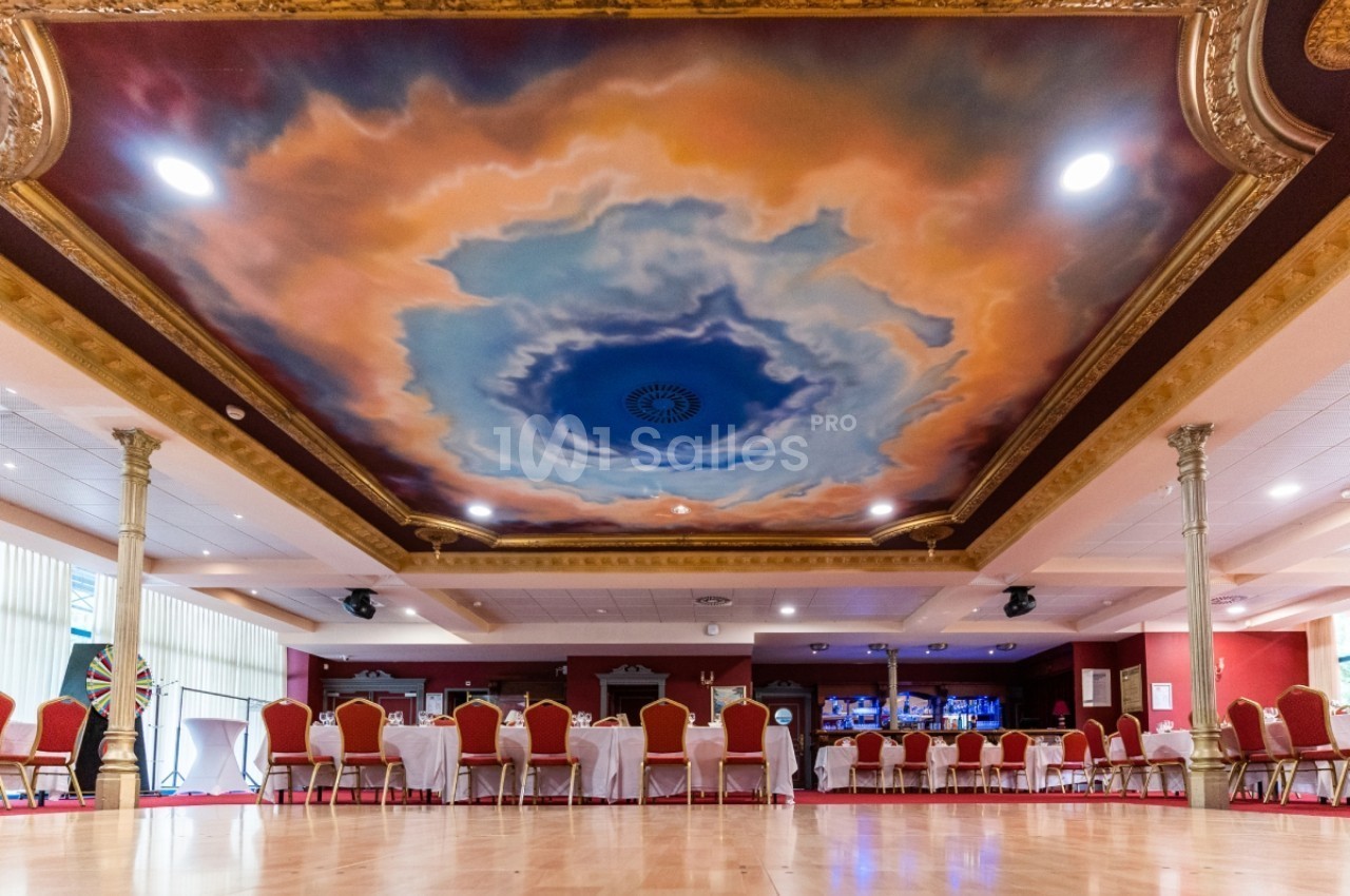 Plafond peint avec un ciel nuageux entouré de moulures dorées dans une grande salle avec des chaises et tables alignées.
