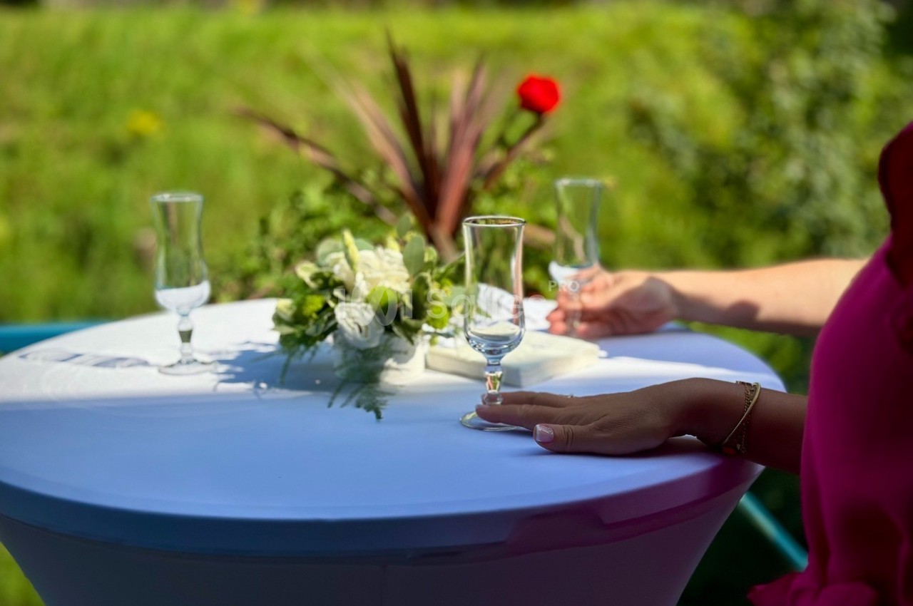 Deux mains posées sur une table haute blanche, décorée de fleurs, avec des verres vides en extérieur verdoyant.
