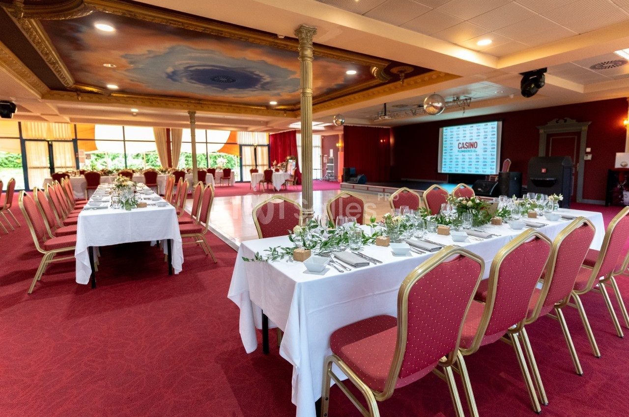 Salle de réception avec tables dressées, nappes blanches, chaises rouges et décoration végétale, prête pour un événement.
