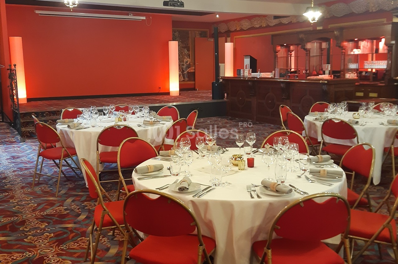 Salle de réception avec tables rondes dressées, chaises rouges et éclairage chaleureux.