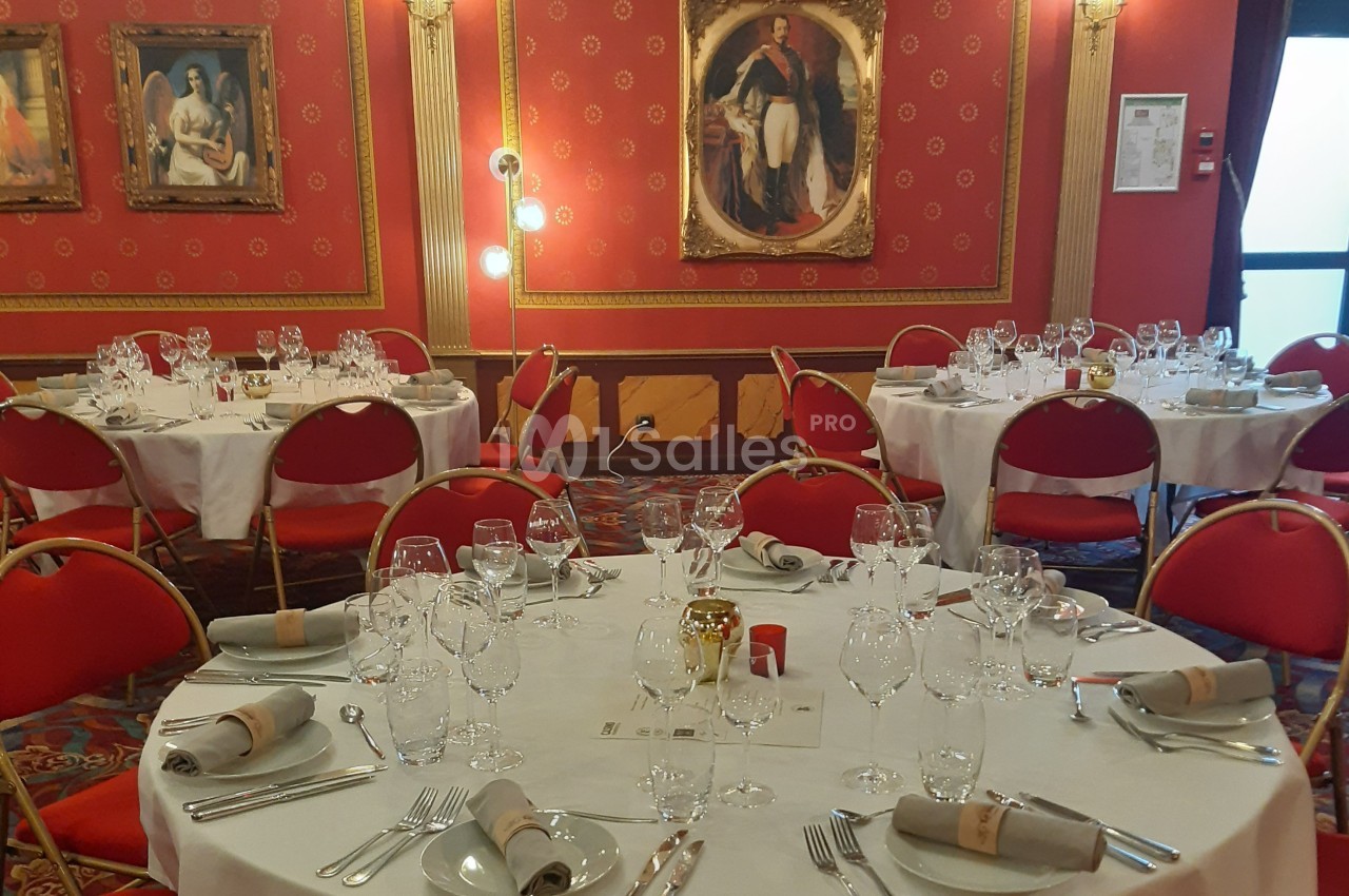 Salle de réception élégante avec tables rondes dressées, nappes blanches, chaises rouges et décorations murales classiques.