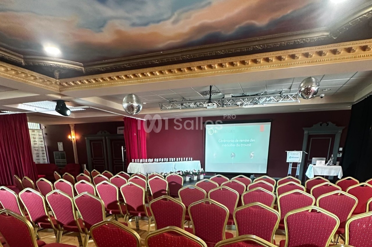 Salle de conférence avec rangées de chaises rouges, écran de projection allumé et décorations au plafond.
