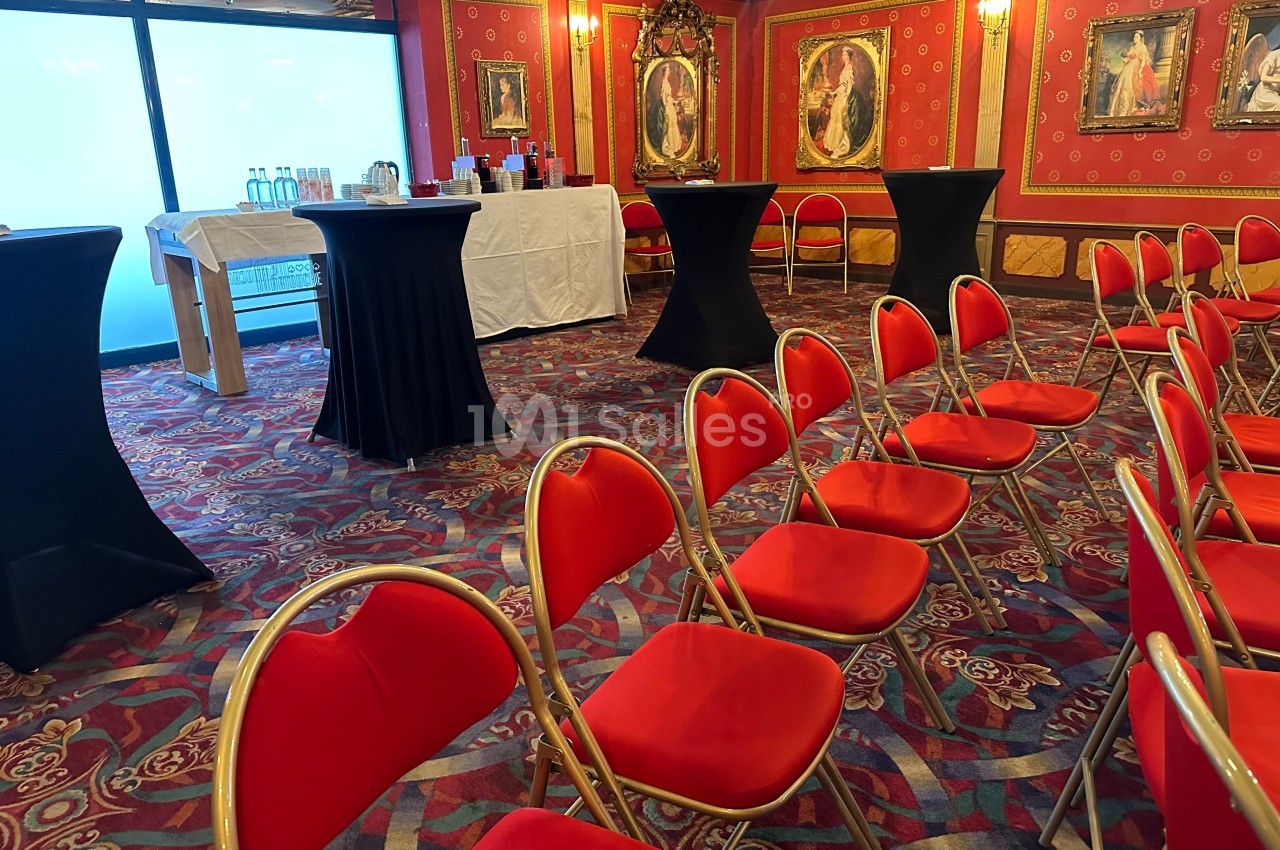 Salle de réception avec chaises rouges alignées, tables hautes noires et buffet en arrière-plan.