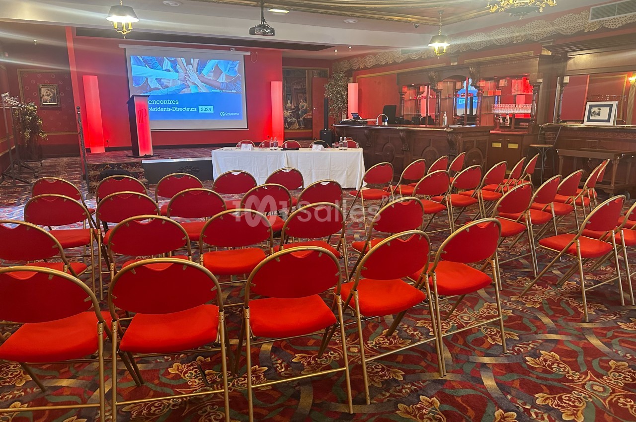 Salle de conférence avec rangées de chaises rouges, écran de présentation allumé et table blanche à l'avant.