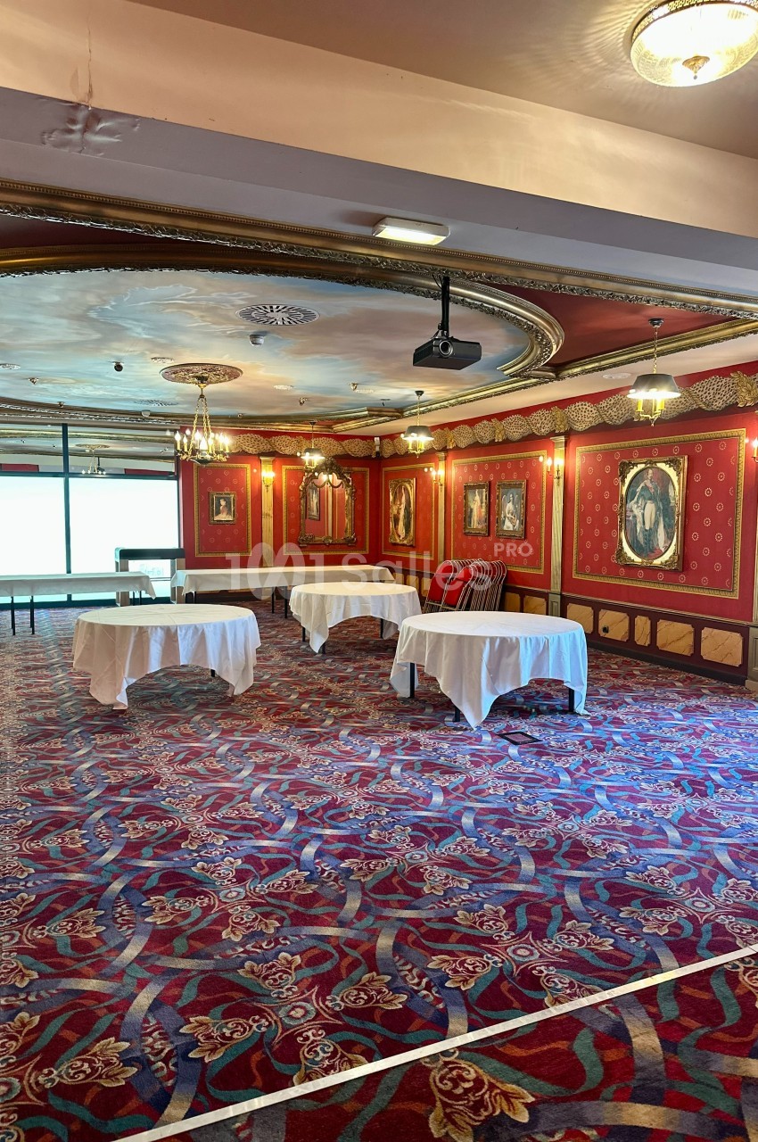Salle décorée de rouge avec tapis à motifs, tables recouvertes de nappes blanches et cadres accrochés aux murs.