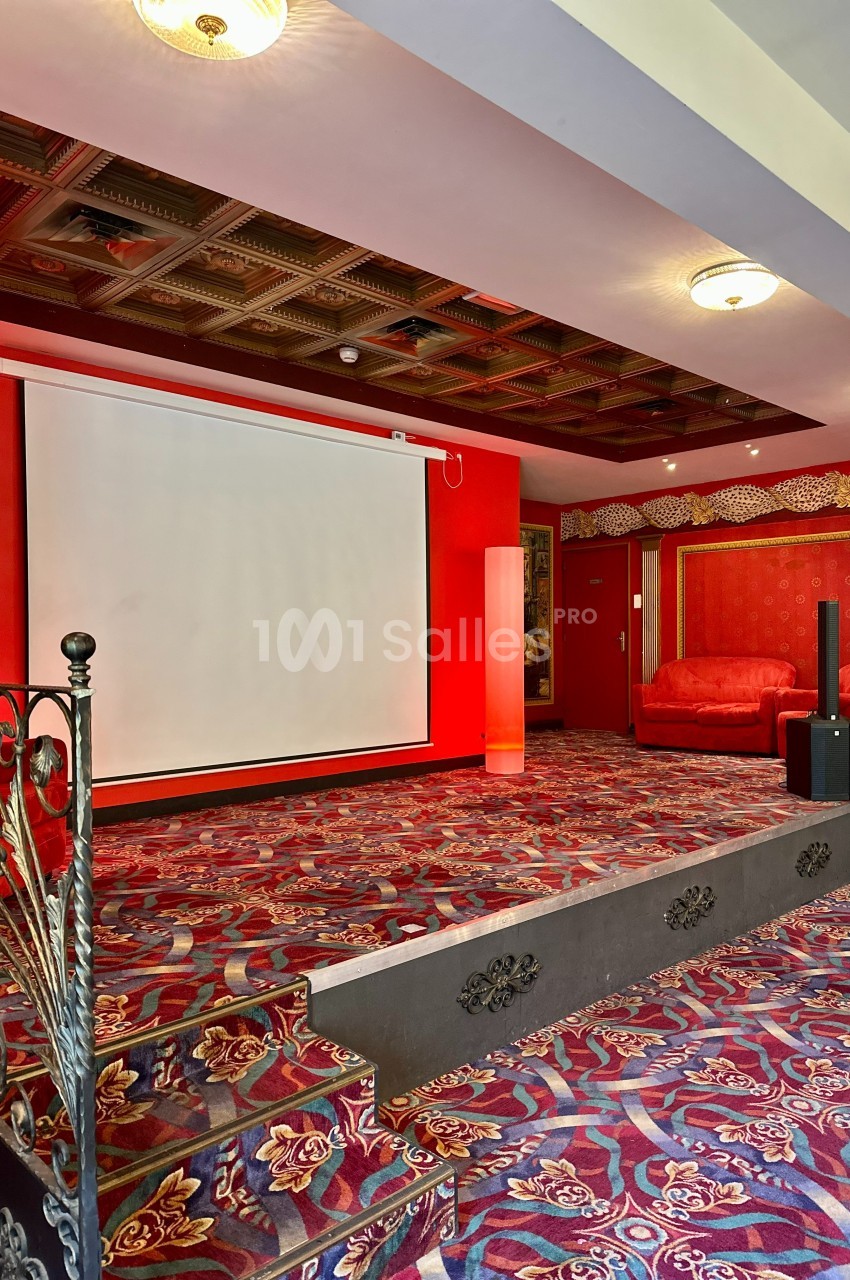 Salle avec écran de projection, tapis rouge à motifs, fauteuils assortis et plafond à caissons décoratifs.