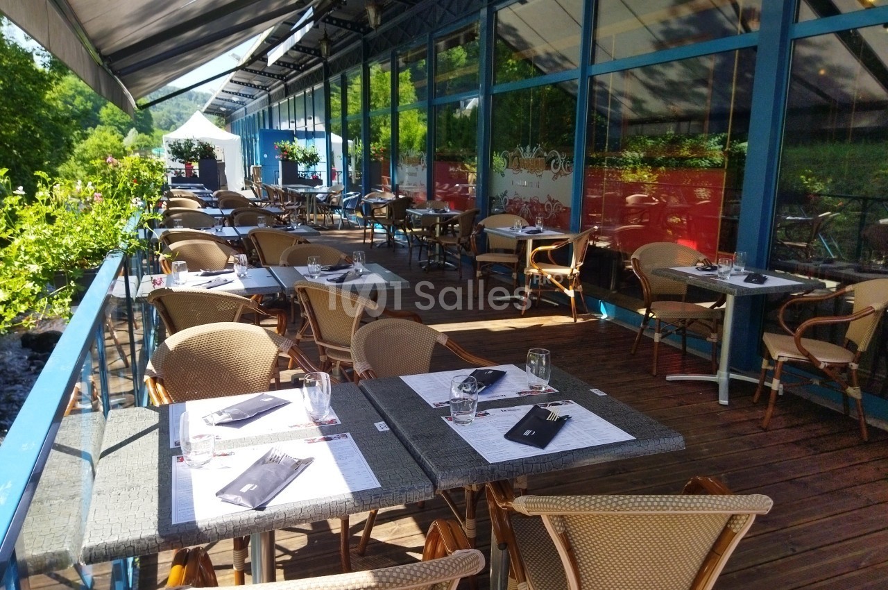 Terrasse ensoleillée d'un restaurant avec des tables dressées, entourée de verdure et bordée par un cours d'eau.