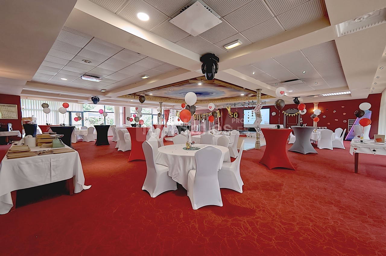 Salle décorée pour un événement avec tables rondes, nappes blanches, ballons colorés et éclairage chaleureux.