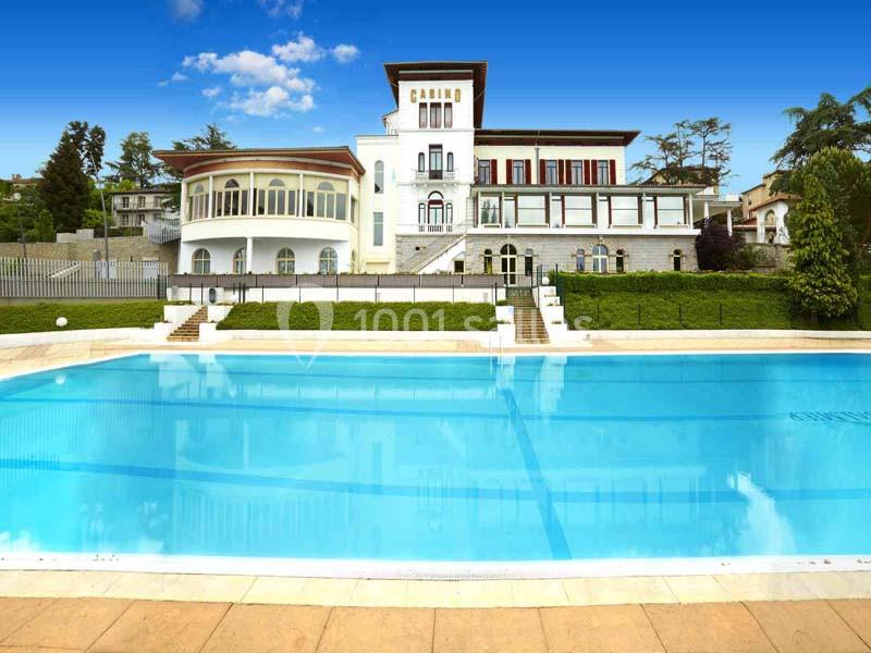 Bâtiment blanc avec balcons et grandes fenêtres, entouré de verdure, devant une piscine extérieure bleue.