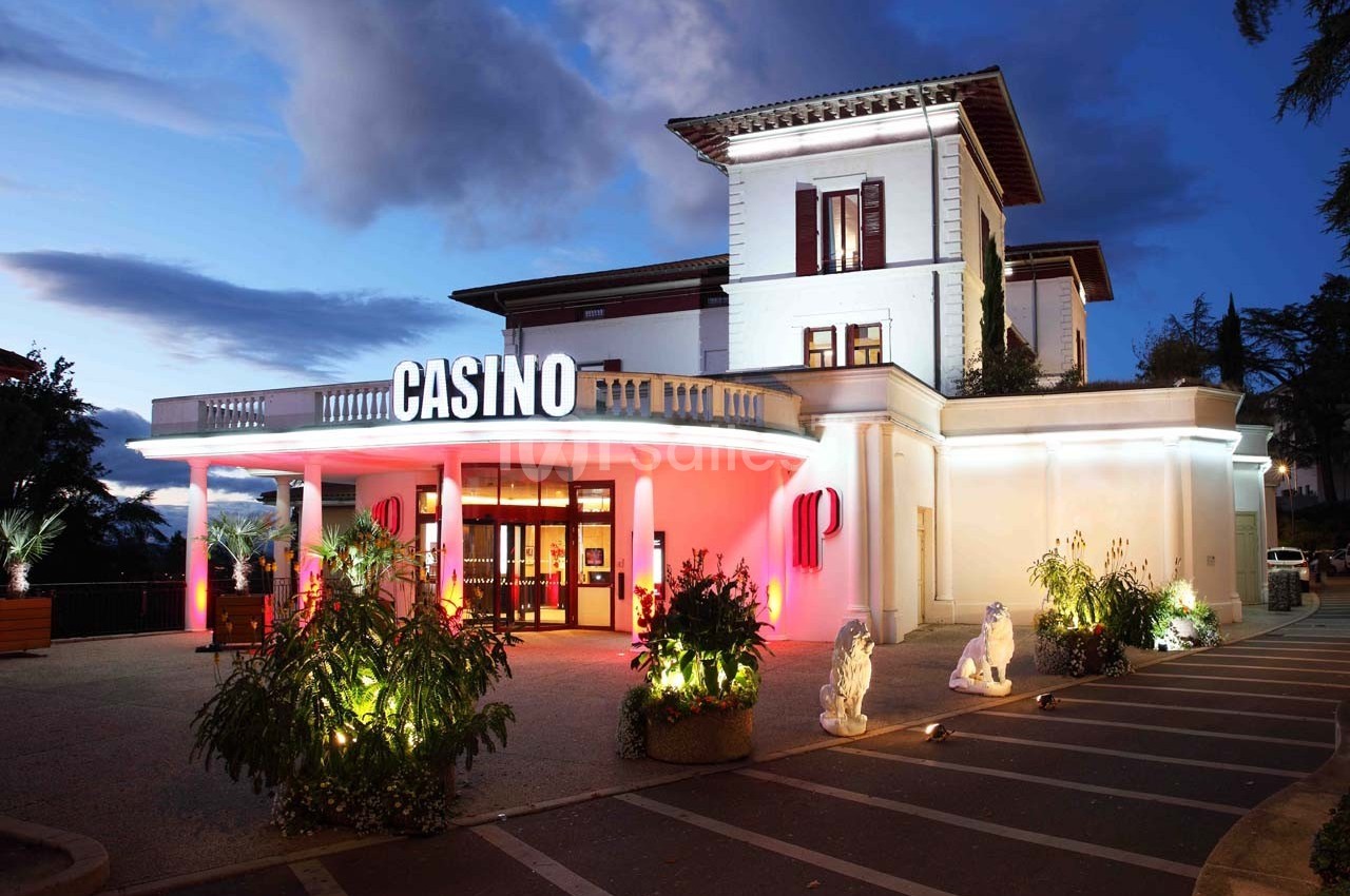 Façade éclairée d'un casino de style classique avec entrée principale entourée de plantes et statues, en soirée.