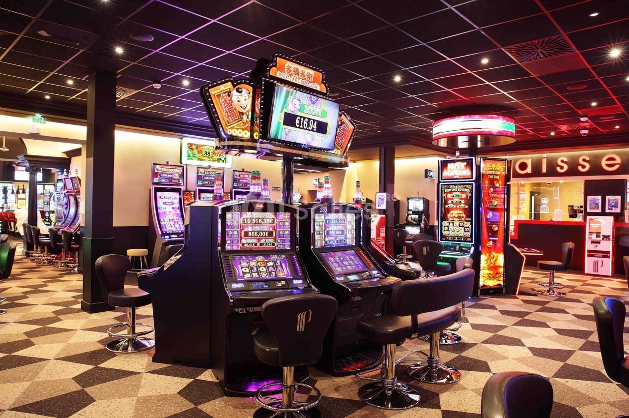 Salle de casino avec machines à sous illuminées, chaises noires et décor moderne sous un plafond sombre.