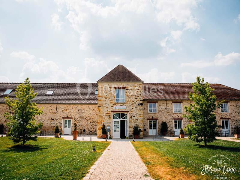 Bâtiment en pierre avec toit en tuiles, entouré de pelouse et d'arbres, sous un ciel partiellement nuageux.
