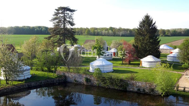 Vue d'un ensemble de yourtes blanches installées dans un cadre verdoyant avec des arbres et un étang au premier plan.