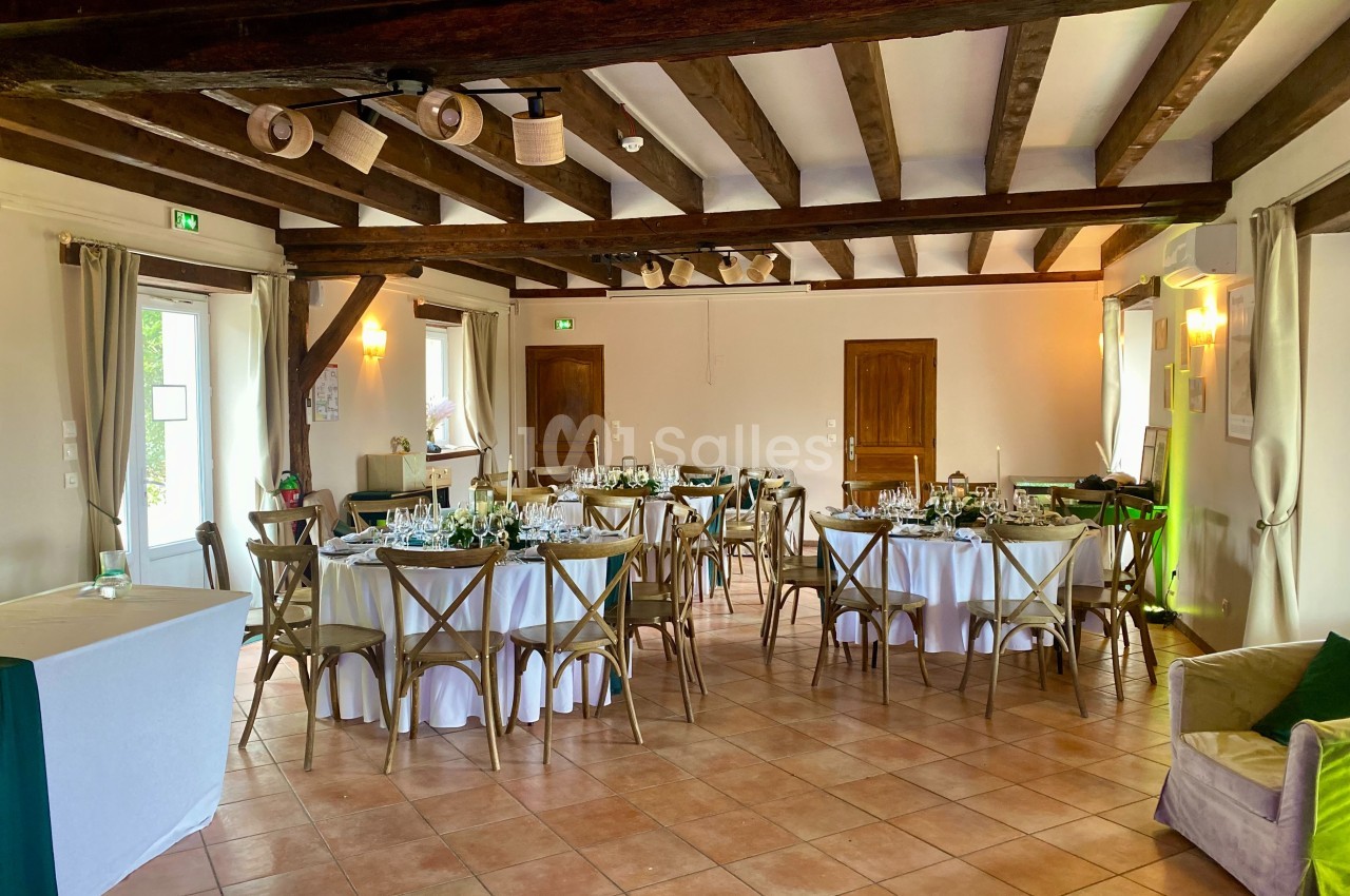 Salle de réception lumineuse avec poutres apparentes, tables rondes dressées et chaises en bois.