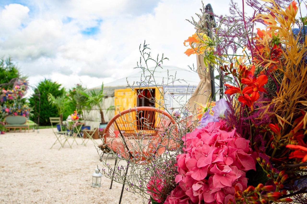 Fleurs colorées au premier plan avec une terrasse extérieure, des chaises et une yourte en arrière-plan sous un ciel nuageux.