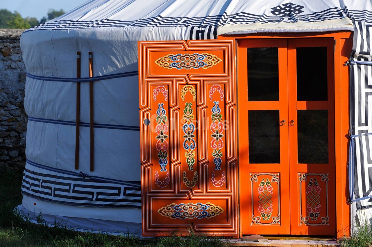 Yourte traditionnelle blanche avec une porte en bois orange ornée de motifs colorés, située en extérieur.