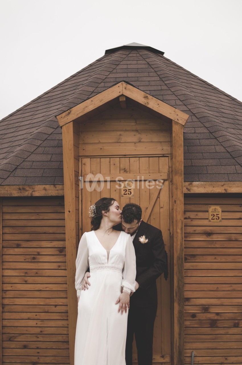 Un couple en tenue de mariage pose devant une petite cabane en bois avec une porte numérotée 25.