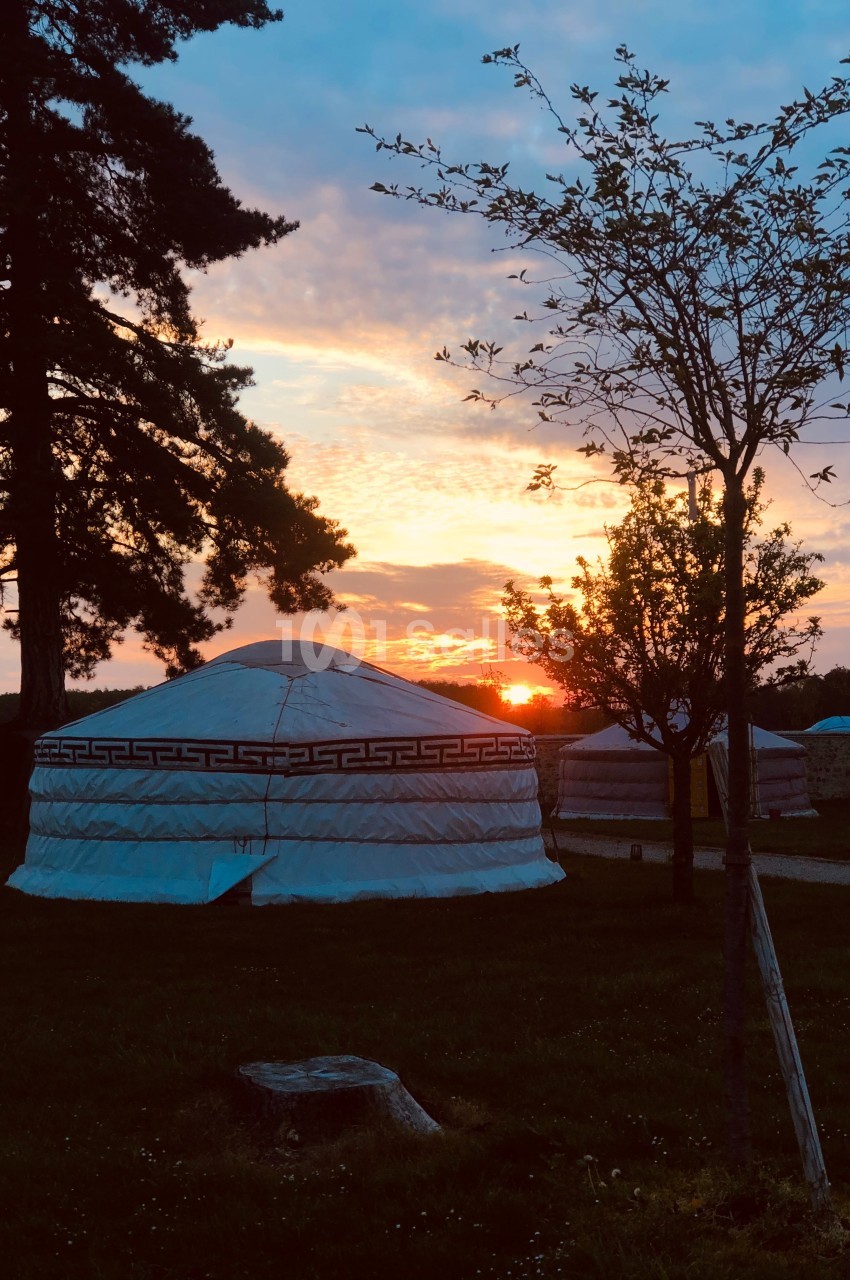 Une yourte blanche dans un paysage au coucher du soleil, entourée d'arbres et d'herbe.