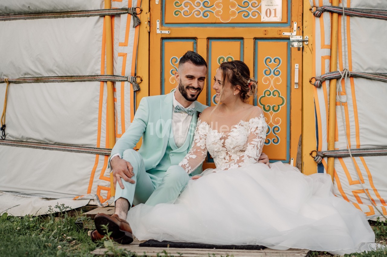 Un couple assis devant une porte colorée d'une yourte, vêtu d'une robe de mariée et d'un costume clair.