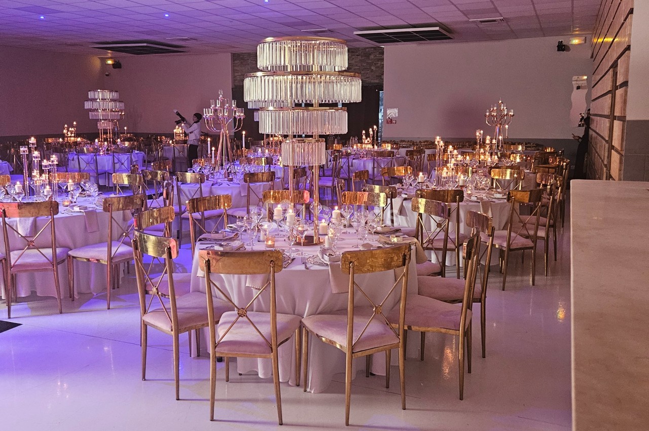 Salle de réception décorée avec des tables rondes, chaises dorées, chandeliers et éclairage tamisé.