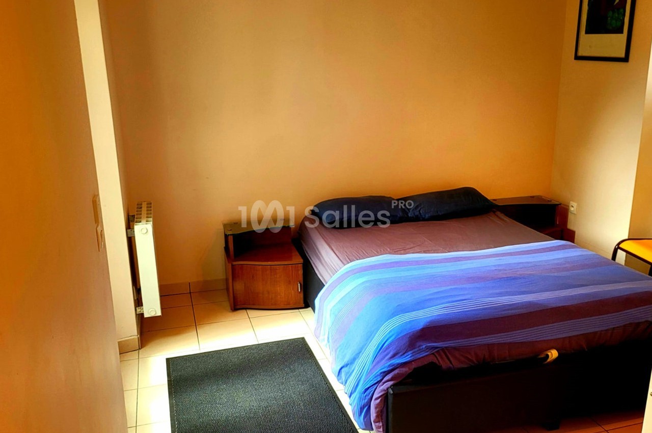 Lit double avec draps bleus dans une chambre simple aux murs clairs, équipée de tables de chevet et d'un radiateur.