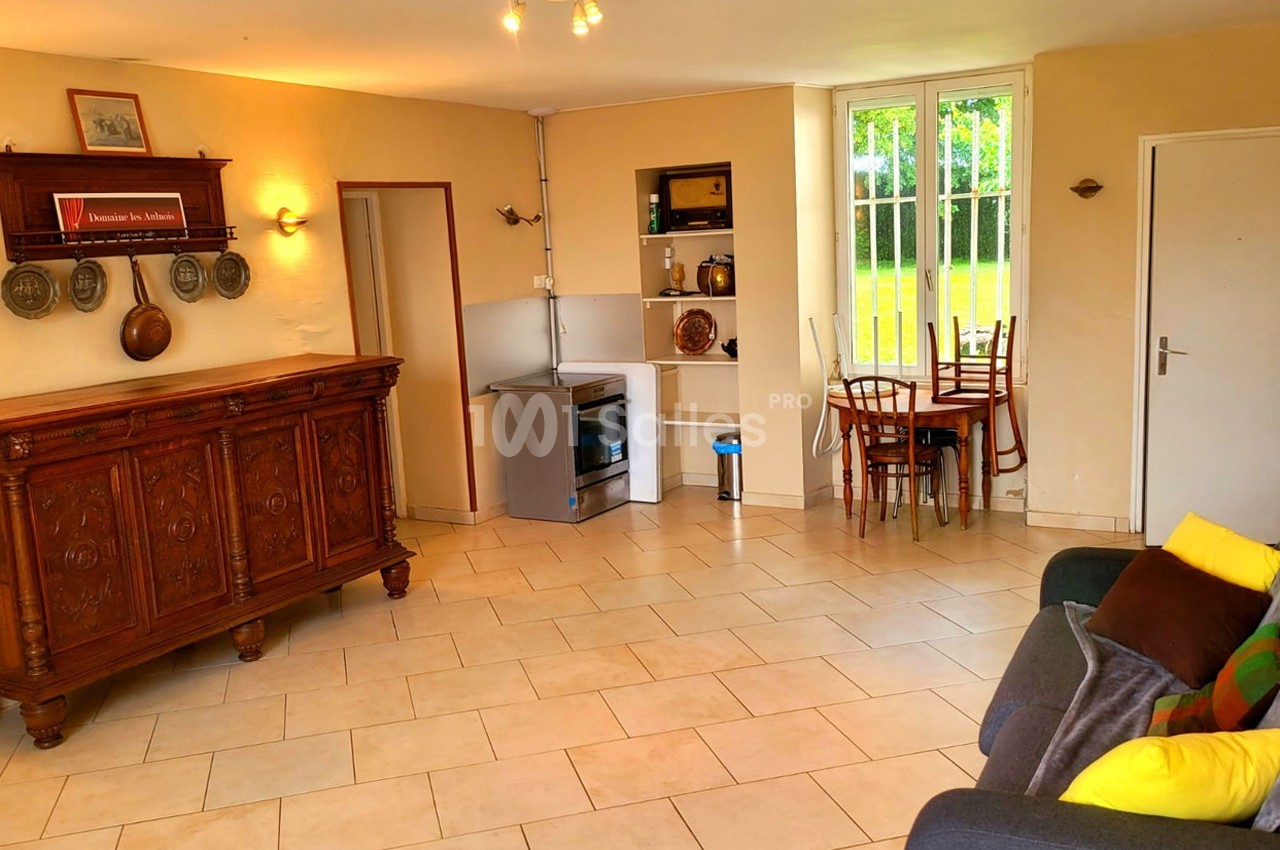 Salon lumineux avec carrelage beige, canapé, meuble ancien, coin cuisine équipé et table près d'une fenêtre.