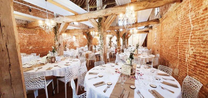 Salle de réception avec murs en briques, poutres apparentes, tables rondes dressées et décorations élégantes.