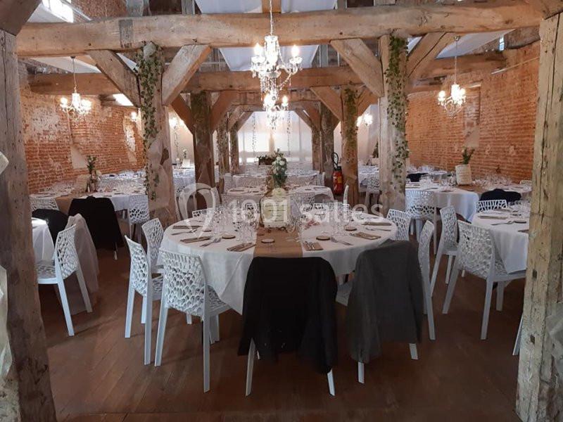 Salle de réception avec murs en briques, poutres apparentes, tables rondes dressées et chaises blanches.