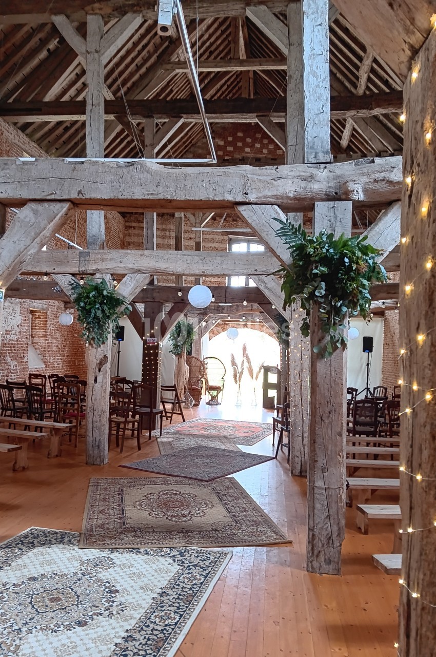 Intérieur d'une grange aménagée avec des poutres en bois, tapis, guirlandes lumineuses et chaises disposées pour un…