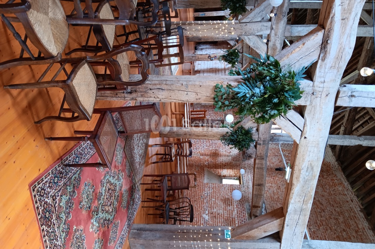 Salle rustique avec poutres apparentes, chaises en bois, tapis coloré et décorations végétales suspendues.