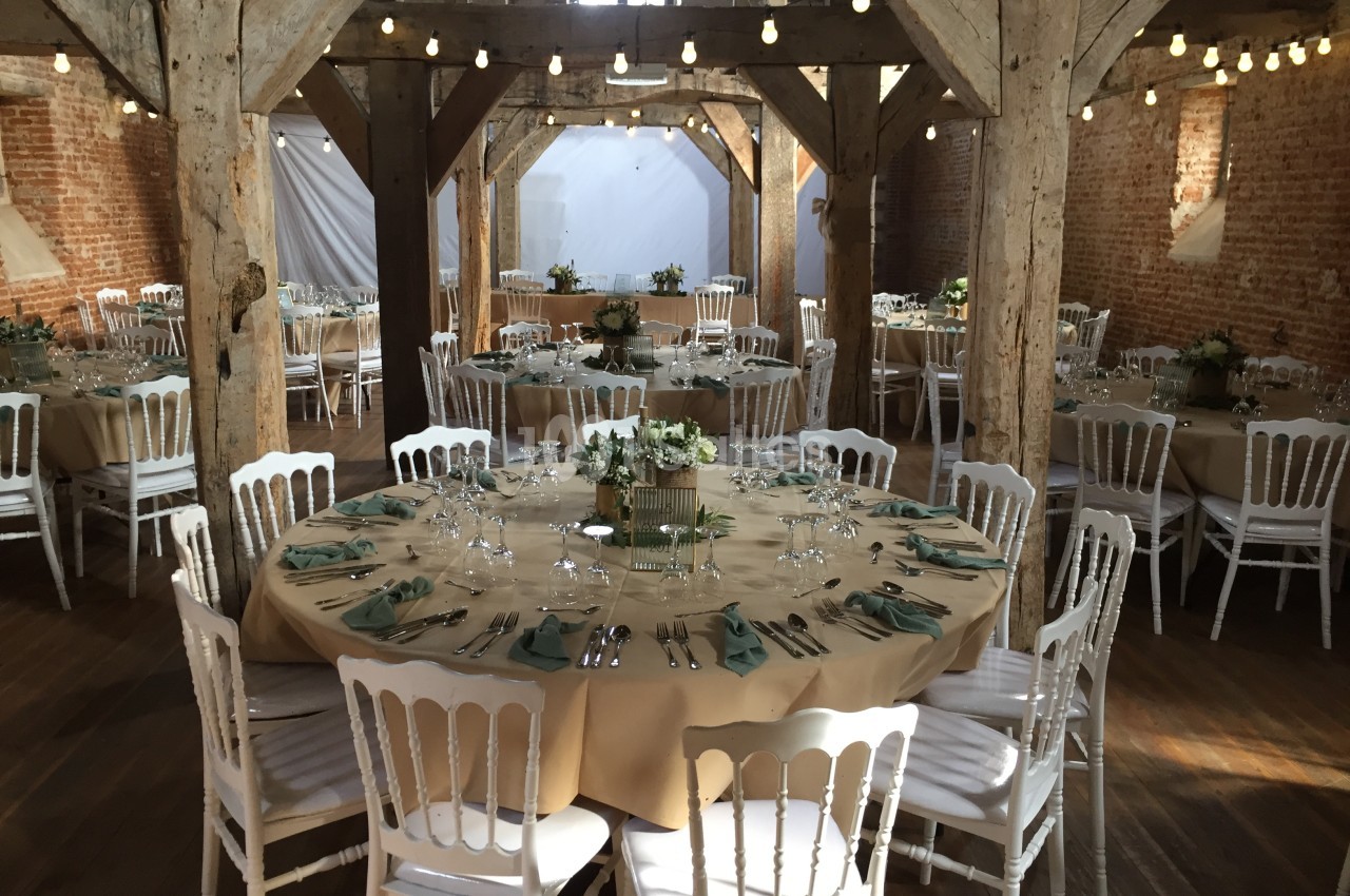 Salle de réception rustique avec tables rondes dressées, nappes beige, chaises blanches et guirlandes lumineuses suspendues.