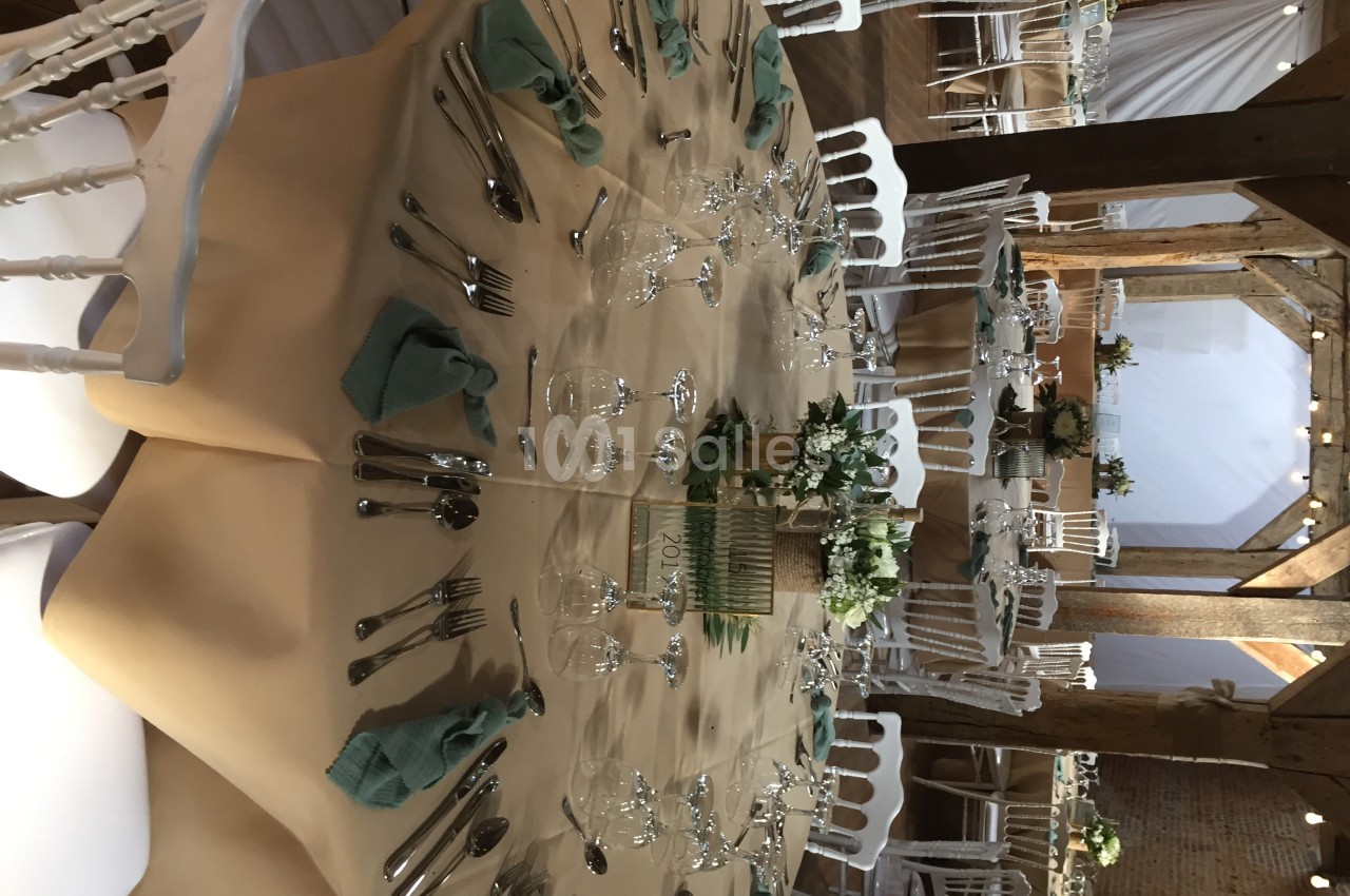 Table ronde dressée avec nappes beige, serviettes vertes, couverts, verres et centre de table floral dans une salle en bois.