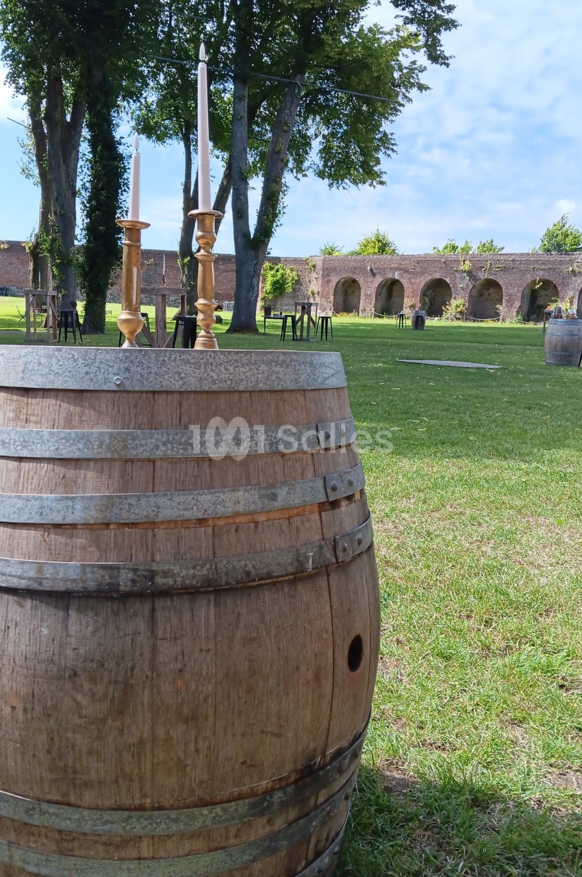 Tonneau en bois utilisé comme table avec des chandeliers, placé dans un jardin verdoyant devant des arches en pierre.