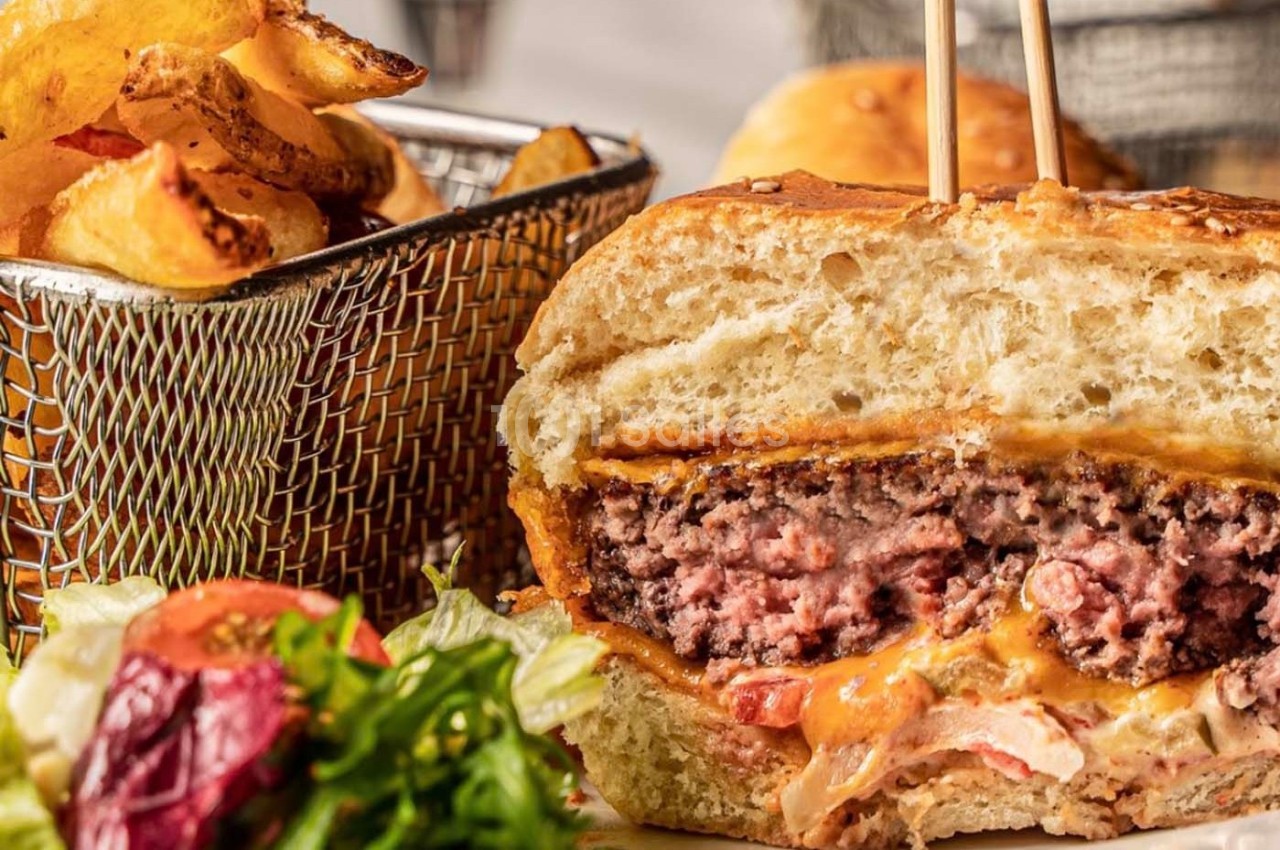 Burger coupé en deux avec fromage fondant, servi avec des frites dorées dans un panier et une salade en accompagnement.