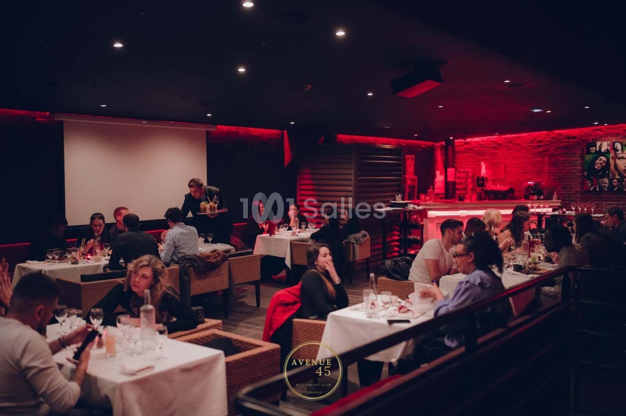 Salle de restaurant avec éclairage tamisé, clients dînant à des tables et un bar en arrière-plan.
