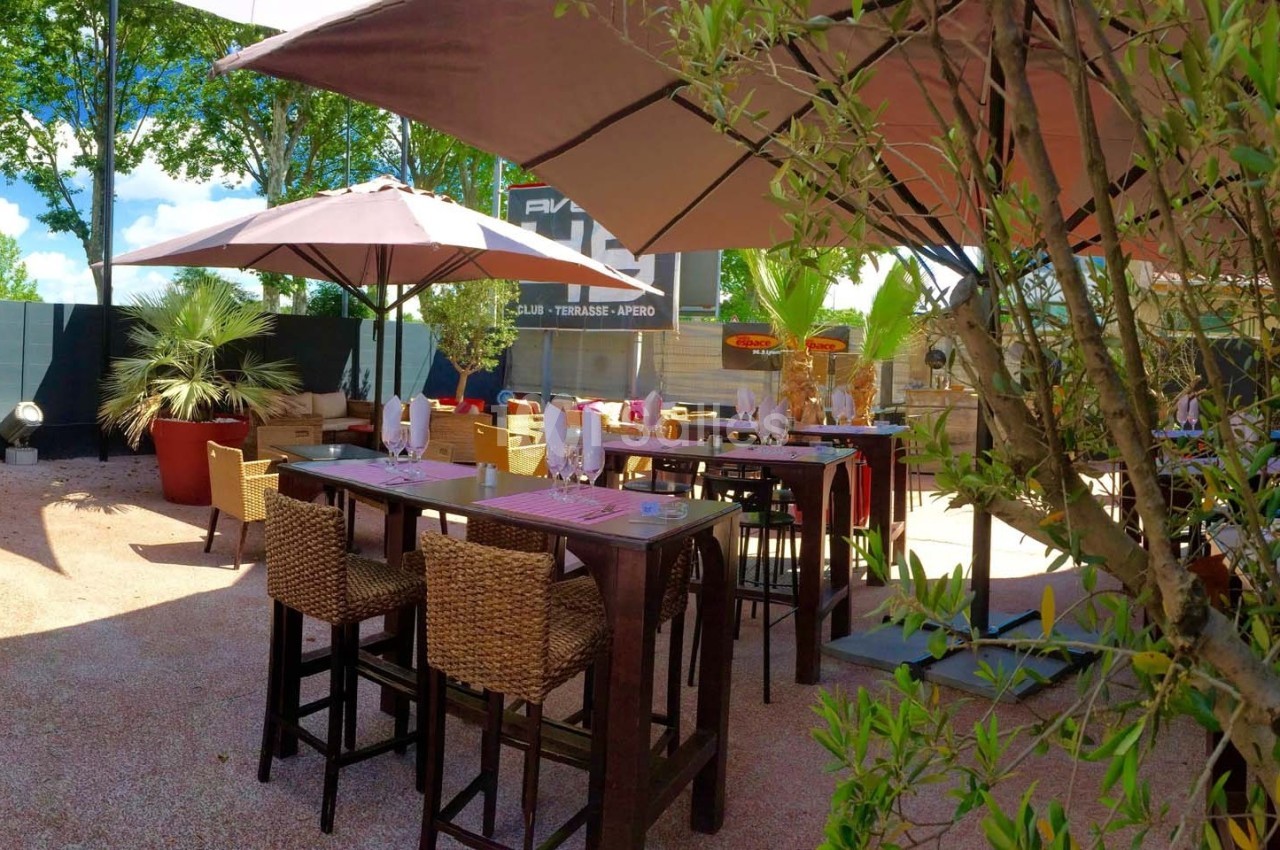 Terrasse extérieure avec tables hautes, chaises en osier, parasols et végétation environnante par temps ensoleillé.