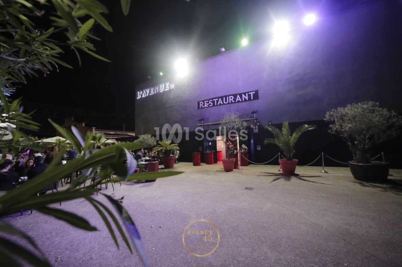 Façade d'un restaurant éclairé la nuit, entourée de plantes en pots et d'un espace extérieur dégagé.