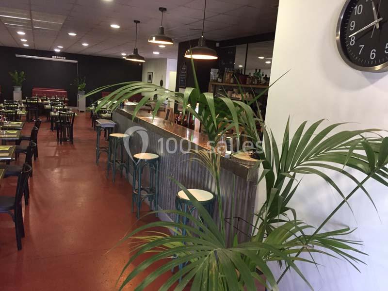 Salle de restaurant avec un long comptoir en métal, des tables dressées et des plantes décoratives au premier plan.