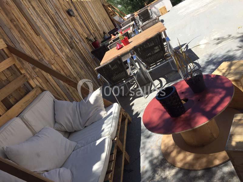 Terrasse extérieure avec canapé, table basse rouge et tables en bois entourées de chaises, ambiance décontractée.