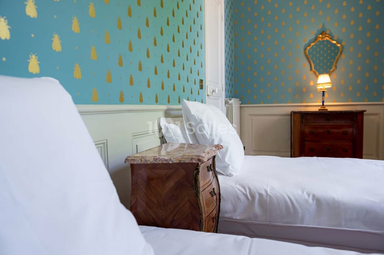 Deux lits simples avec table de chevet en bois, mur décoré de motifs dorés et miroir au-dessus d'une commode.