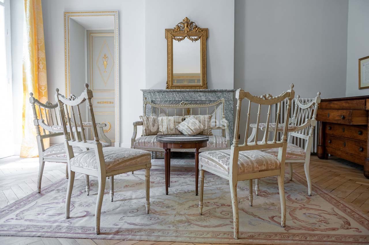 Mobilier ancien avec chaises et table en bois clair, disposés autour d'un tapis, devant une cheminée et un miroir doré.